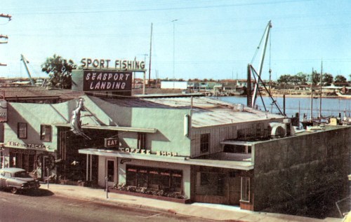 A vintage view of Seaport Landing with a sport fishing sign, shops, and boats in the background by the water.