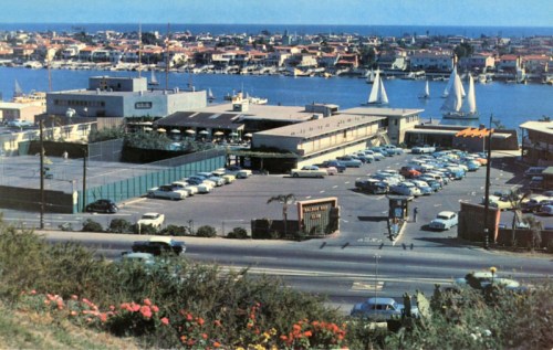 Aerial view of a marina with sailboats, a parking lot, and buildings along the waterfront. Bright blue sky and water.