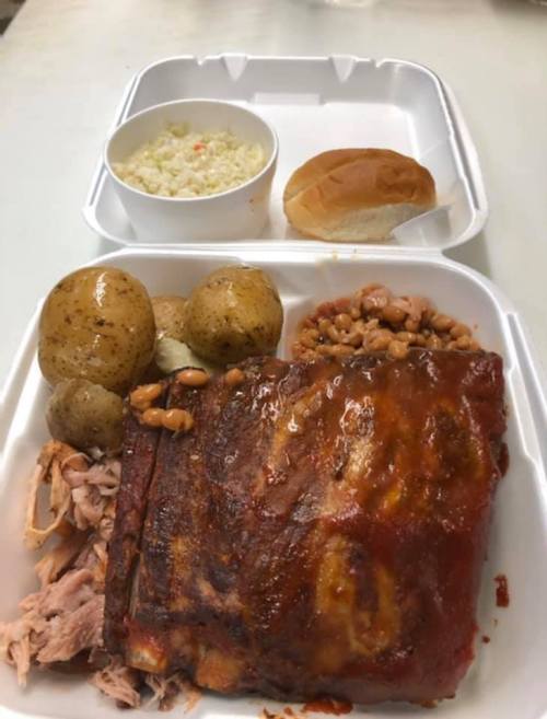 A takeout container with barbecue meat, baked potatoes, baked beans, coleslaw, and a roll.