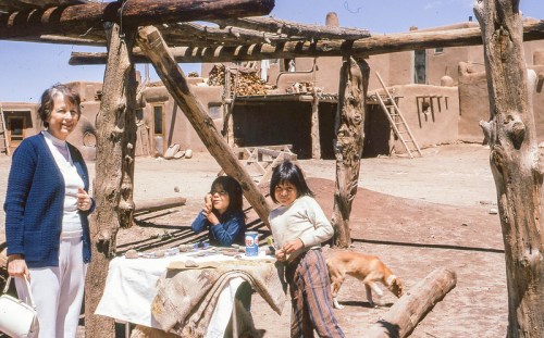 A woman and two girls sit at a table outside a rustic adobe structure, with a dog nearby and a clear blue sky above.