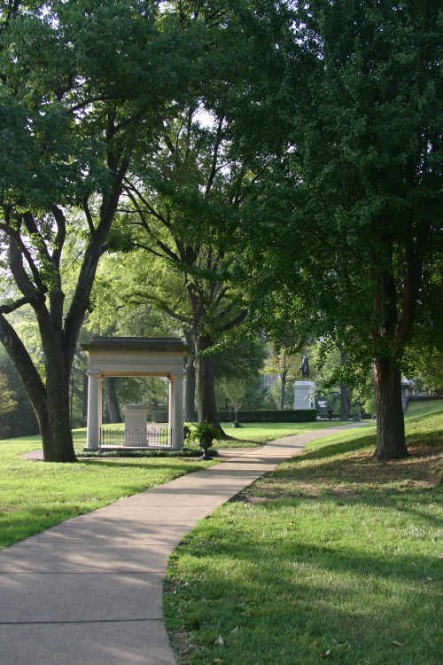 A winding path through a green park, lined with trees and a gazebo, leading to a distant statue.