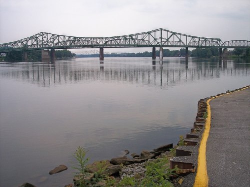 A green bridge spans a calm river, reflecting the cloudy sky, with a rocky shoreline and yellow-painted edge in the foreground.