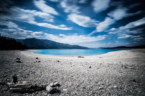 A serene lake with a pebbly shore, surrounded by mountains and a blue sky with scattered clouds.