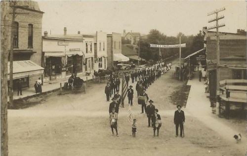 A historic black-and-white photo of a parade with people marching down a street, featuring buildings and a horse-drawn carriage.
