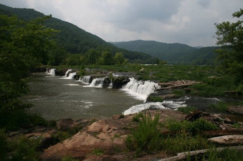 A serene landscape featuring a waterfall cascading over rocks, surrounded by lush greenery and mountains in the background.