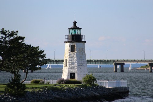 A white lighthouse stands by the water, with a bridge in the background and sailboats in the distance under a clear sky.