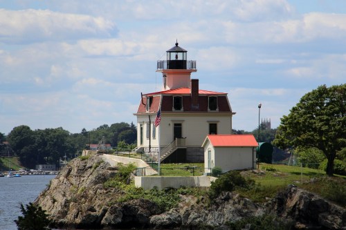 A white lighthouse with a red roof stands on a rocky shore, surrounded by water and trees under a blue sky.