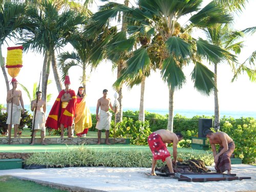 Two men prepare a traditional feast outdoors, surrounded by palm trees and onlookers in colorful attire.