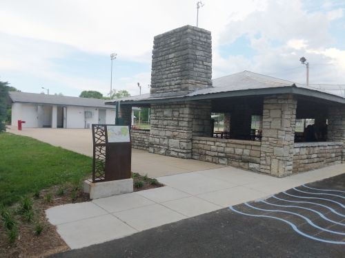 A stone pavilion with a chimney, next to a paved area and restrooms, surrounded by green grass and trees.