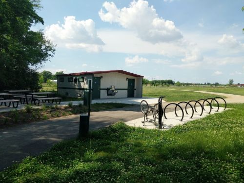 A small building with a red roof, bike racks, and picnic tables in a grassy area under a blue sky with clouds.