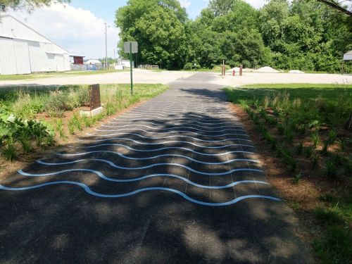 A paved path with blue wave patterns, surrounded by greenery and trees, under a bright sky.