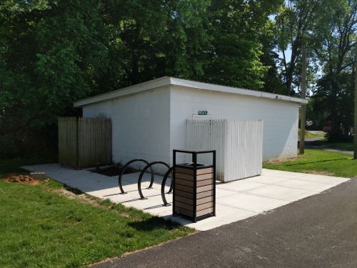 A white building with a wooden fence, bike racks, and a trash bin on a paved area surrounded by grass and trees.