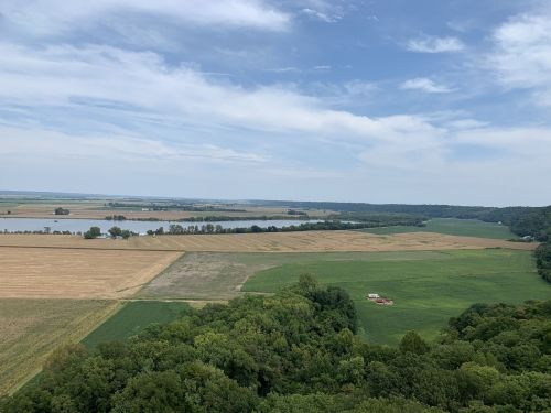 A panoramic view of fields and forests under a blue sky, with a river visible in the distance.
