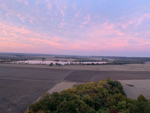 A scenic view of farmland and a river at sunset, with colorful clouds and trees in the foreground.