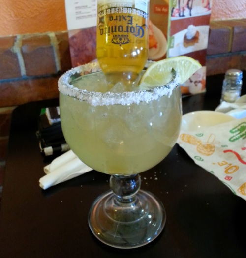 A large margarita glass with a salted rim, ice, a lime wedge, and a beer bottle perched on top.