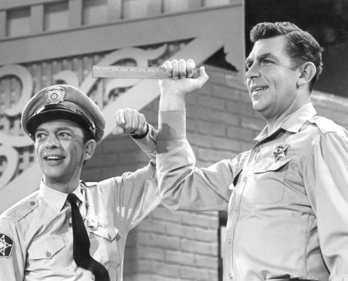 Two smiling police officers hold up a ruler, posing playfully in a black-and-white scene.