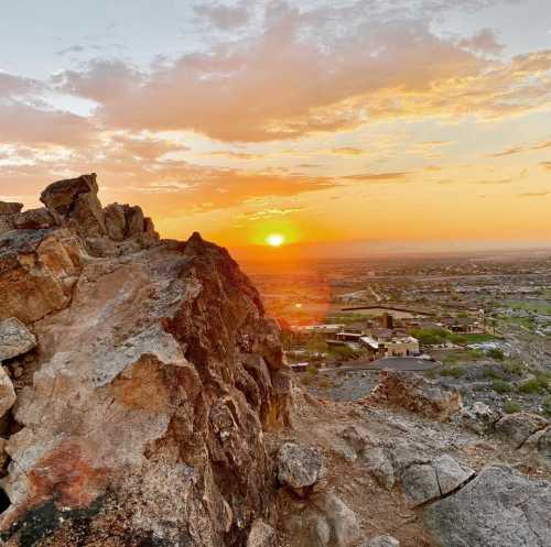 A rocky landscape at sunset, with vibrant orange and yellow hues illuminating the sky and distant hills.