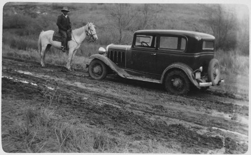 A man on a horse stands beside a vintage car on a muddy road, with a rural landscape in the background.