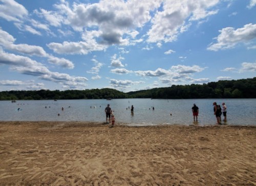 A sandy beach with people swimming and playing in a lake under a partly cloudy sky.