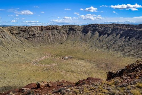 A vast, dry crater surrounded by rocky terrain under a blue sky with scattered clouds.