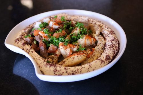 A plate of hummus topped with grilled meat, tomatoes, and parsley, served in a curved white dish.