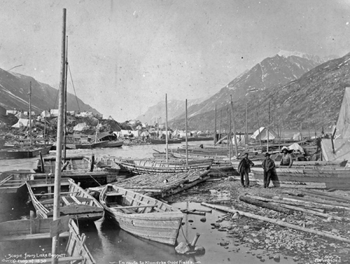 Black and white photo of a lakeside scene with boats, mountains, and two figures walking along the shore.
