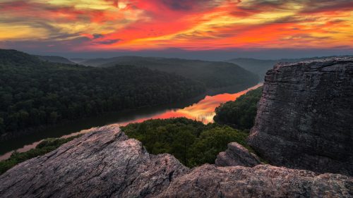 A vibrant sunset over a river, surrounded by lush green hills and rocky cliffs, creating a serene landscape.