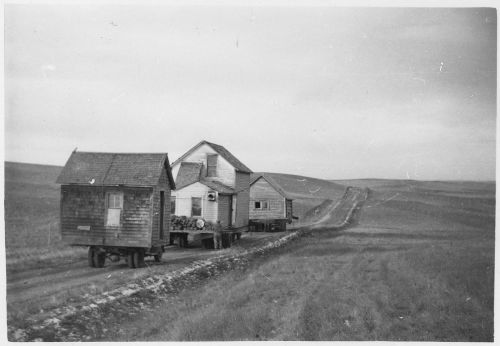 Three wooden houses on wheels traveling down a dirt road through a vast, open landscape. Black and white photo.