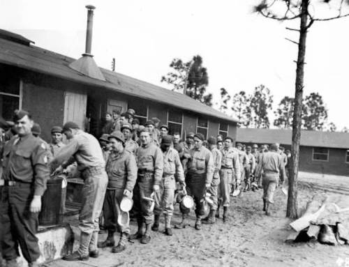 Black and white photo of soldiers in line outside a building, holding plates, likely waiting for a meal.