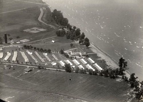 Aerial view of a lakeside camp with tents, fields, and trees along the shoreline.
