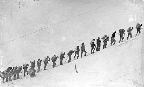A line of climbers ascending a snowy slope, carrying gear, in a black and white photograph.