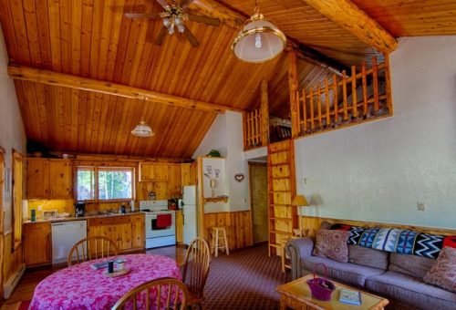 Cozy cabin interior with wooden walls, a kitchen, dining area, and a lofted sleeping space. Warm and inviting atmosphere.