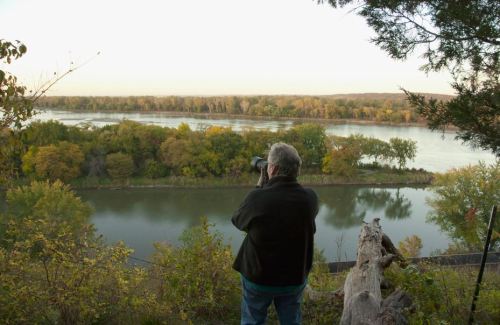 A person stands by a river, photographing the scenic landscape with trees and autumn colors in the background.