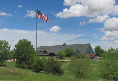 A large American flag flies in front of a green-roofed building surrounded by trees and a blue sky with clouds.