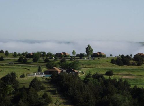 A scenic view of green hills with houses, surrounded by fog and trees under a clear sky.