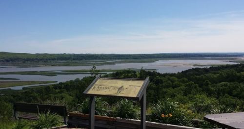 Scenic view of a river and lush greenery, with an informational sign in the foreground under a clear blue sky.