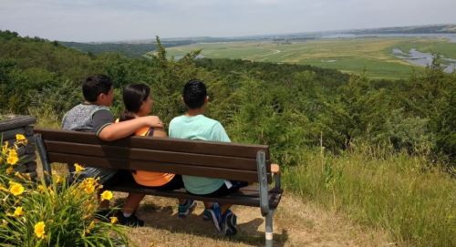 Three children sit on a bench, overlooking a scenic landscape with greenery and a river in the distance.