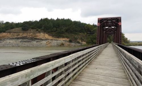 A wooden bridge leads towards a rocky hillside, with trees lining the riverbank under a cloudy sky.