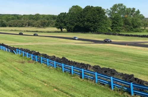 Cars racing on a track surrounded by green grass and a blue tire barrier under a clear sky.