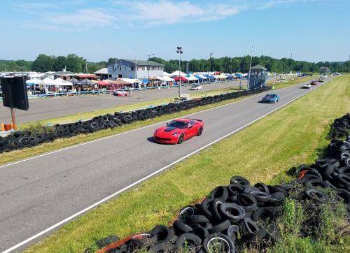 A red sports car races on a track lined with tires, with tents and spectators in the background under a clear blue sky.