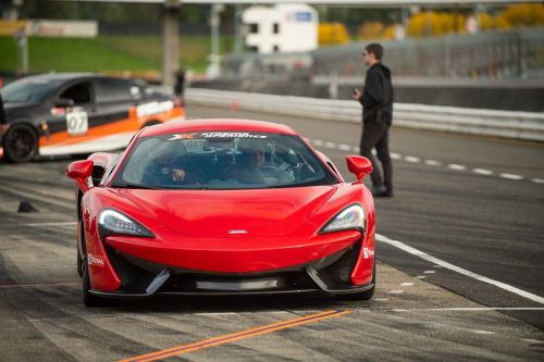 A red sports car on a racetrack, with a blurred background of another car and a person standing nearby.