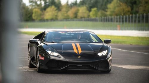 A sleek black sports car with orange stripes drives on a racetrack surrounded by greenery.