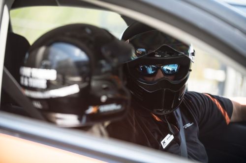 A driver in a racing helmet and sunglasses sits in a car, with another helmet visible in the foreground.