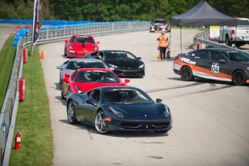 A line of sports cars, including a black Ferrari and a red Lamborghini, on a racetrack with spectators in the background.