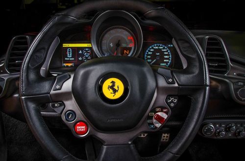 Close-up of a Ferrari steering wheel with a yellow logo, dashboard displays, and engine start button.