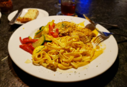 A plate of fettuccine pasta with grilled chicken, mixed vegetables, and a slice of garlic bread in the background.