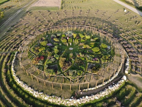 Aerial view of a circular garden with intricate flower beds and wooden structures, surrounded by rows of plants.