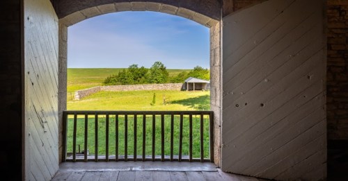View from an arched window overlooking a green field and a stone wall under a clear blue sky.