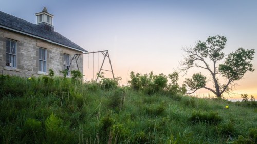 A weathered building beside a swing set, with tall grass and a lone tree against a colorful sunset sky.