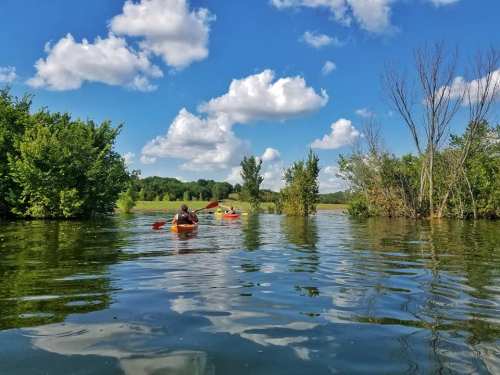 Two people kayaking on a calm lake surrounded by lush greenery and fluffy clouds in a blue sky.